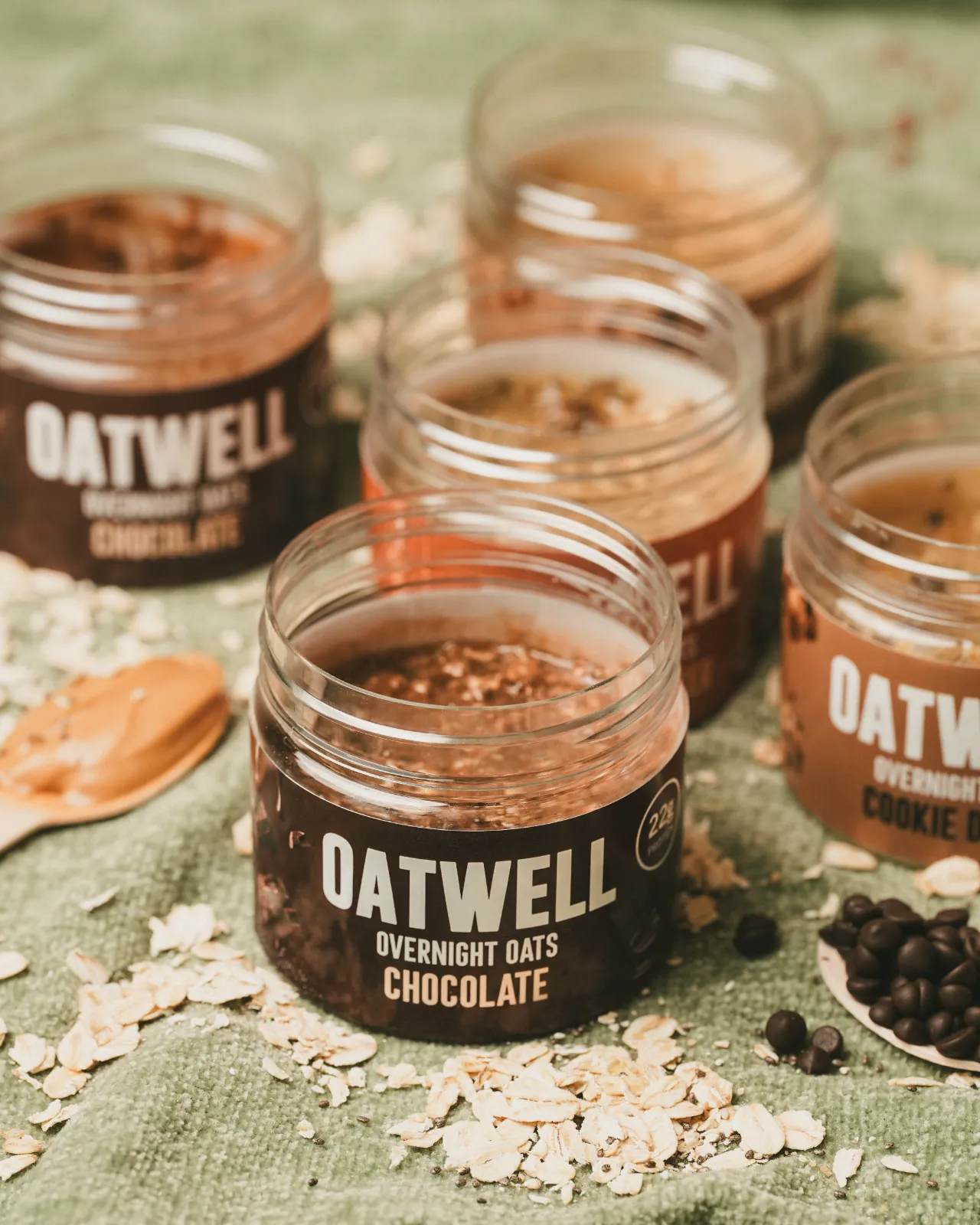 Oatwell jars open with rolled oats and chocolate chips on green linen
